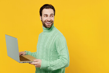 Young smiling happy freelancer programmer man 20s wearing mint knitted sweater hold use work on laptop pc computer look aside on workspace area isolated on plain yellow background studio portrait