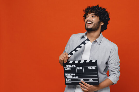 Overjoyed excited laughing jubilant stunning young bearded Indian man 20s years old wears blue shirt holding classic black film making clapperboard isolated on plain orange background studio portrait.