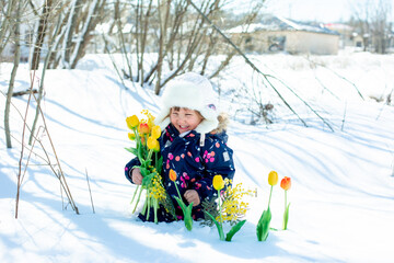 funny little girl in overalls and a hat in winter holds a bouquet of flowers. The concept of spring...