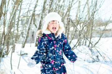 a funny little girl in overalls and a hat catches snow with her tongue in winter outside