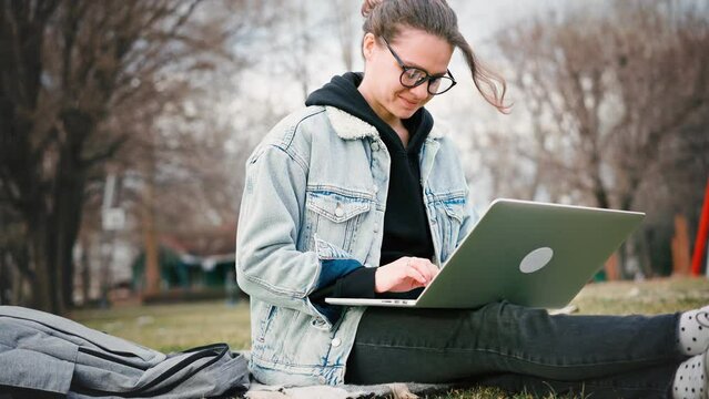 Young woman student wearing glasses typing on her laptop while sitting on the lawn in the park.