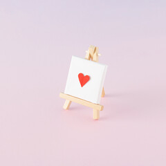 Creative arrangement made of easel and white canvas with a red heart on a pink background. Minimal love concept.