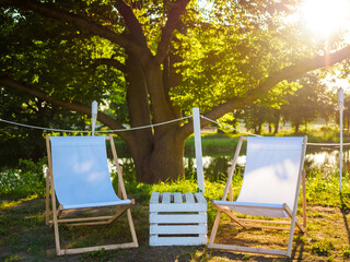 two white beach seat chairs and wooden box as a table between it on nature on a lake shore, city break and place for relax