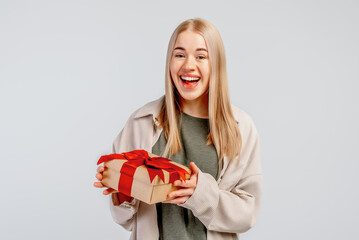 Image of Happy young blonde woman in shirt holding gift box with red ribbon and looking at the camera over gray background - Powered by Adobe