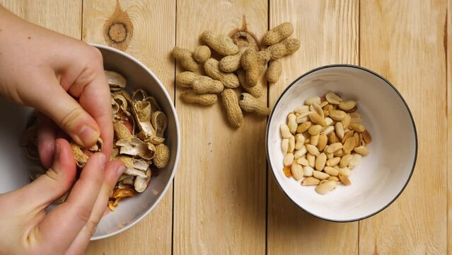 Slow motion footage of a woman's fingers peeling peanuts from their shells. A handful of peanuts lie on a wooden table. View from above. Peeling an earthy peanut at home