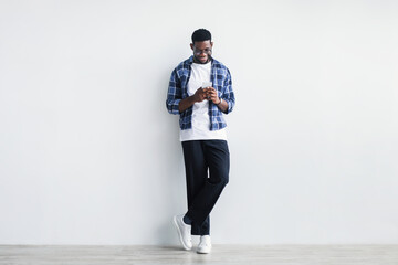 Joyful young black guy using modern smartphone, working remotely or video chatting, standing...