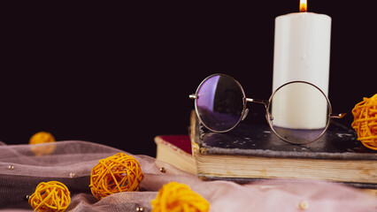 Burning candle books and thin veil with decor. Composition of burning wax candle near eyeglasses on stack of books near thin fabric with decorative wicker balls on black background