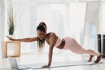 Fit young black woman standing in plank with outstretched arm, following online video tutorial on...