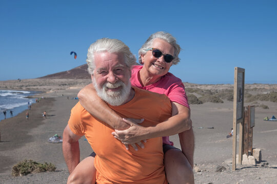 Portrait of happy mature bearded man at the beach carrying piggyback his elderly wife. Senior caucasian couple smiling enjoying sunlight and sea during travel vacation or retirement