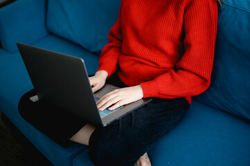 Naklejka premium a young girl in a red knitted sweater works on a laptop at home. she is sitting on a blue sofa