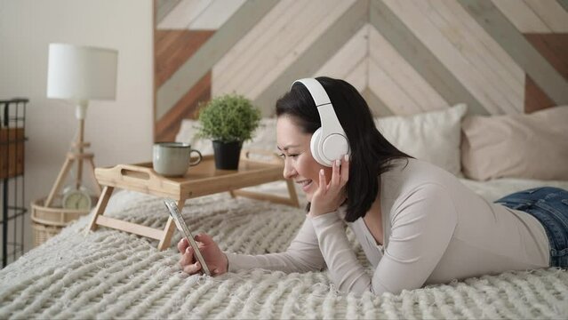 Young asian ethnic woman using smart phone surfing social media, checking news, playing mobile games or texting messages sitting on sofa. Calm girl spending time at home with gadget technology.