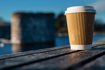 Cardboard cup with plastic lid for coffee or tee on a wooden bench in a park. Blue cloudy sky in the background and river with remains of an old bridge. Outdoor refreshment concept
