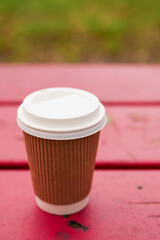 Cardboard cup with plastic lid for coffee or tee on a wooden red bench in a park. Outdoor refreshment concept. Selective focus.
