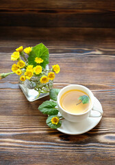 Fresh spring coltsfoot flowers and tea cup on wooden table. Healing herbal infusion with medicinal plant Coltsfoot (Tussilago farfara). 