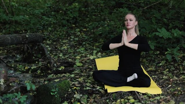 Beautiful middle aged woman practice yoga in autumn forest near river. Female seats on yellow yoga mat on ground in covered by dry leaves woodland and doing exercises.
