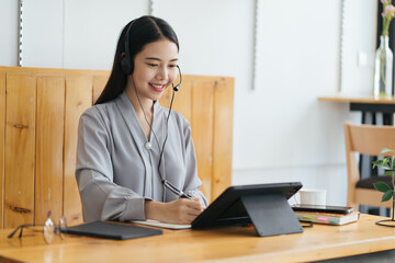 Cheerful beautiful Asian woman wearing headphones and look to digital tablet at video calling meeting and study online on the Internet.