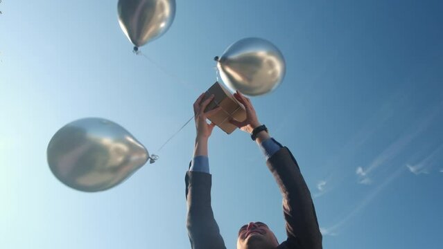 Man catches grey box attached to silver helium balloons against blue sky. Airdrop of inflatable balls falling down in hands. Low angle shot super slow motion