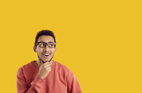 Cheerful curious student thinking about something. Happy positive young ethnic man in casual sweatshirt and glasses looks up at yellow colour text copyspace background for interesting question or idea