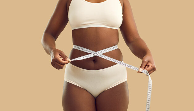 Close up crop of fat African American woman in underwear on yellow studio background measure waist with tape. Overweight black girl make measurement before diet. Weight loss and body shape.
