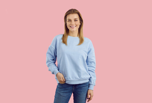 Young model posing in studio. Studio shot of cheerful woman in casual outfit. Happy smiling teen girl wearing pale blue sweatshirt and jeans standing isolated on solid pastel pink colour background
