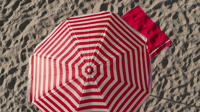 Striped red-white umbrella and towel put on soft sand on sunny day. Waves of sea wash shoreline. Beach essentials on resort in summer aerial view