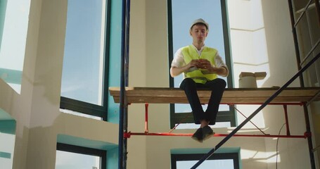 A worker eats a sandwich on the construction site, sits high on scaffolding
