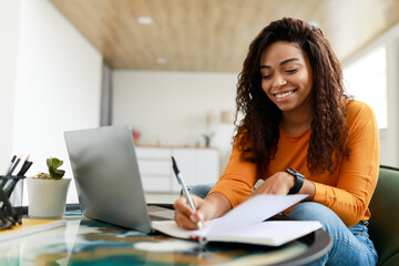 Fototapeta Black woman sitting at desk, using computer writing in notebook obraz