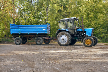 A blue agricultural tractor with a trailer on a dirt road, against a backdrop of bushes with green leaves. Special equipment for different types of work