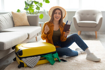 Happy young woman with passport and tickets sitting near suitcase and making YES gesture at home