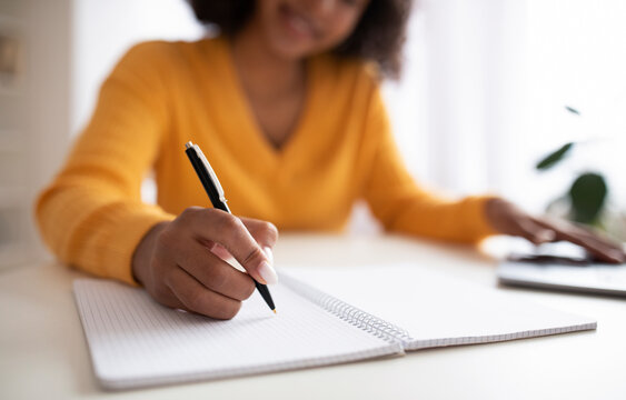 Unrecognizable young black woman writing down info in notebook during online lesson on laptop from home, closeup - Powered by Adobe
