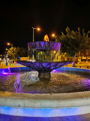 A purple-lit fountain on a central street against a dark sky.