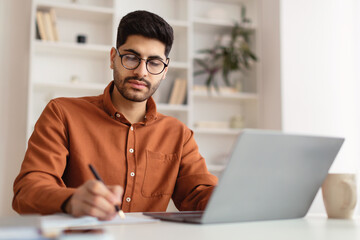 Focused Arab man in glasses using laptop and writing