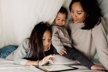 Happy asian family using tablet computer while sitting on bed under blanket