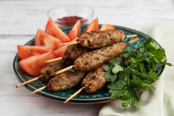 Blurred image of lula kebab on a plate with greens and tomato slices on a light table and adjika in a gravy boat.