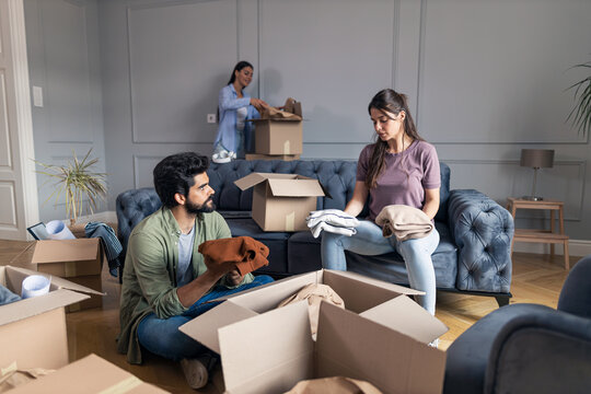 Man and woman in new apartment unpack their clothes from cardboard box while friend helps them.