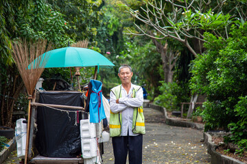 Portrait happy old Asian man street cleaner standing with his arms crossed next to an old gabage cart before going to work with nature background. - obrazy, fototapety, plakaty