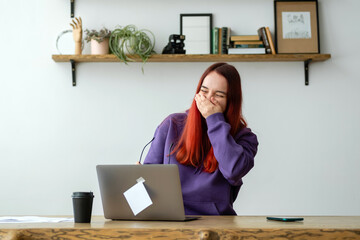 Modern woman in purple hoodie and glasses looks  at laptop screen and laughs very hard sitting at a...