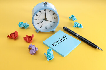 Lifestyle and people concept. Alarm clock, message on What's Next on the blue notepad, pen, and crumpled papers on the yellow background