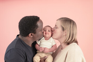 mom and dad give a kiss to the newborn - closeup portrait of a modern multi-ethnic family over pink background