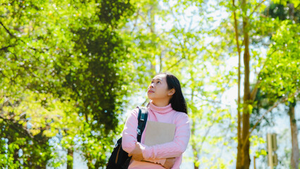 Asian female student with backpack holding a book and looking away in summer park.