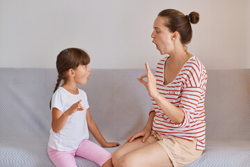 Side view of woman teacher wearing striped shirt and shorts sitting on sofa with little girl, preschooler kid practicing correct pronunciation with a female speech therapist. - obrazy, fototapety, plakaty