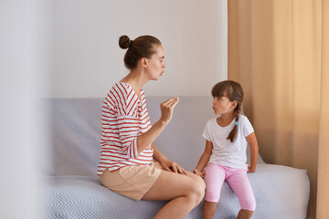 Side view of woman with bun hairstyle wearing striped shirt and shorts sitting on sofa with daughter, preschooler girl practicing correct pronunciation with a female speech therapist.