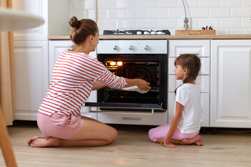 Back view of mother and daughter wearing casual style clothing, woman taking out pie from gas oven,...
