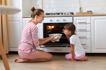 Indoor shot of smiling happy family, mother and daughter sitting on floor near oven and smelling tasty dessert, expressing positive emotions, cooking together, preparing sugary pie.