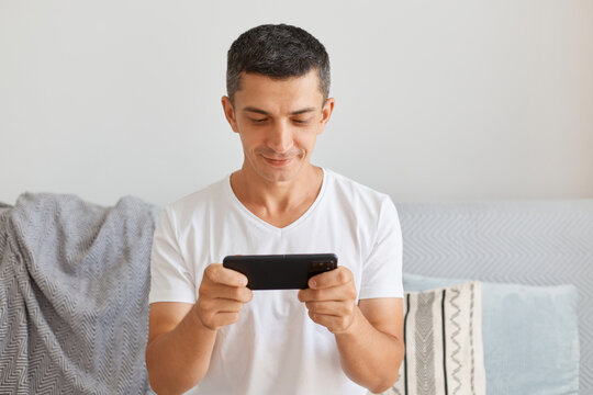 Image of smile positive man wearing casual style white t shirt sitting on cough and holding smart phone in hands, looking at display with optimistic expression, playing video game.