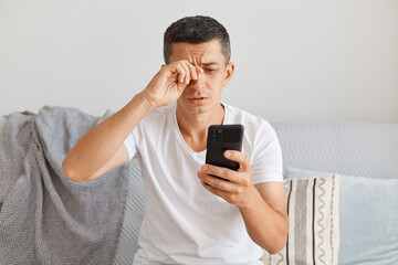 Portrait of tired upset man wearing casual style white t shirt sitting on cough, using smart phone, looks exhausted, rubbing eye, looking at gadget screen with sorrow.