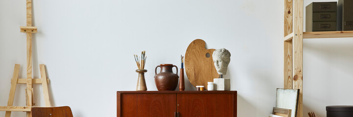 Stylish composition of artist workspace with design retro teak commode, chair, bookcase, poster frames, easel, decoration and elegant personal accessories. Template.