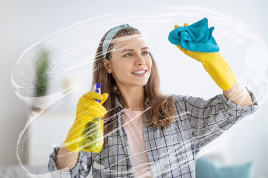 Smiling young woman cleaning soap sud on window with rag, using detergent to wash glass, indoors
