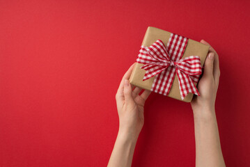 First person top view photo of saint valentine's day decorations young woman's hands demonstrating kraft paper giftbox with checkered ribbon bow on isolated red background with copyspace
