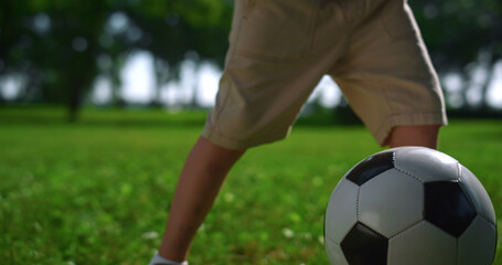 Closeup kid feet kicking up football ball. Boy play on green lawn in park.